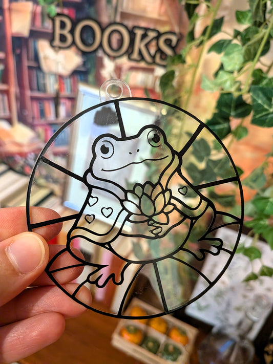 Unbemalter runder DIY-Sonnenfänger mit Frosch Valentin und Seerosenblüte, frontal vor einem Bücherregal in der Hand gehalten.