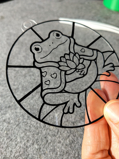 Noch nicht ausgemalter runder DIY-Sonnenfänger mit Frosch Valentin, Cape und Seerosenblüte, in der Hand gehalten