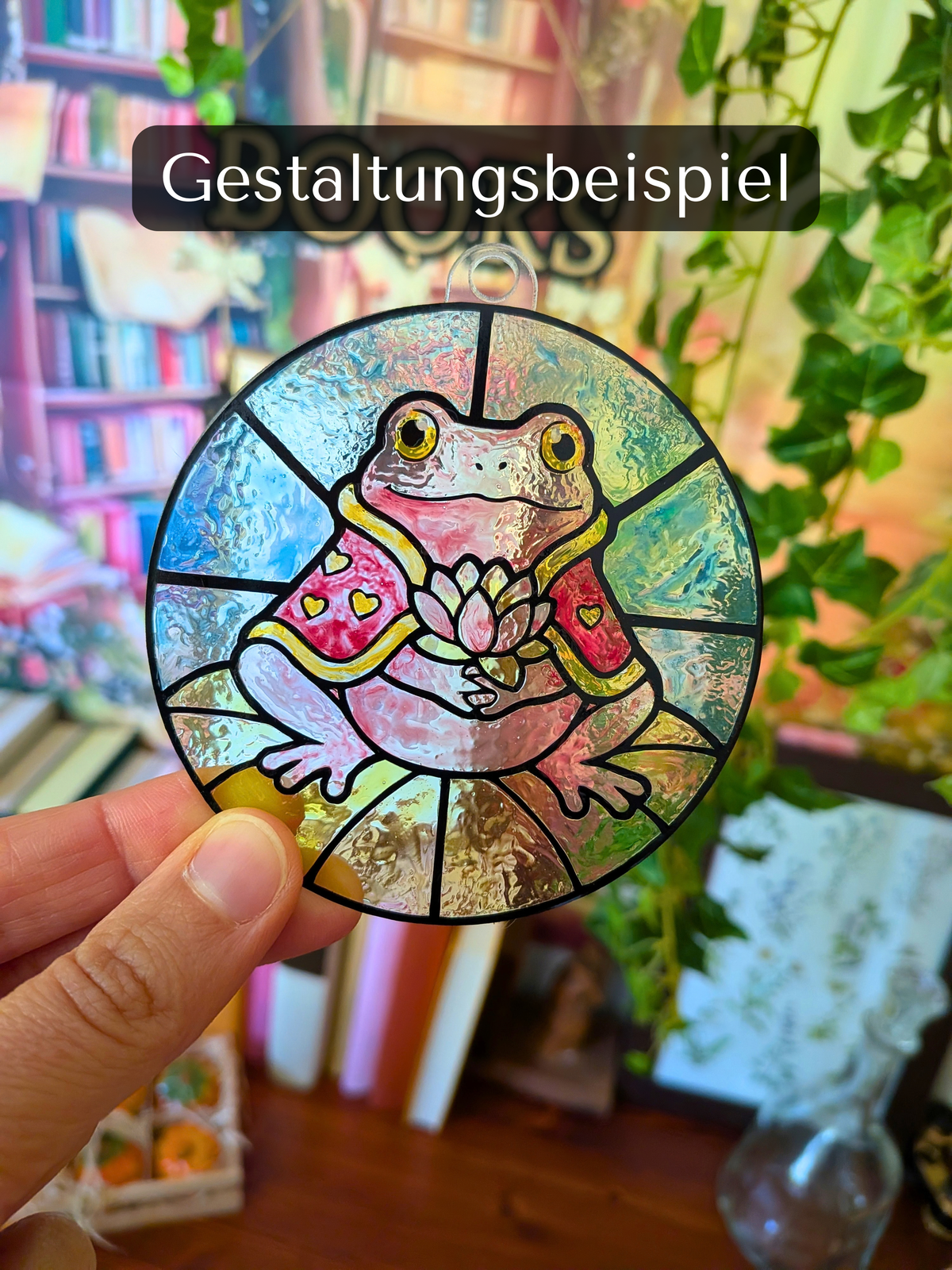 Ausgemalter DIY-Sonnenfänger mit Frosch Valentin in Rosa-, Gelb- und Blautönen, vor einem Bücherregal in der Hand gehalten.